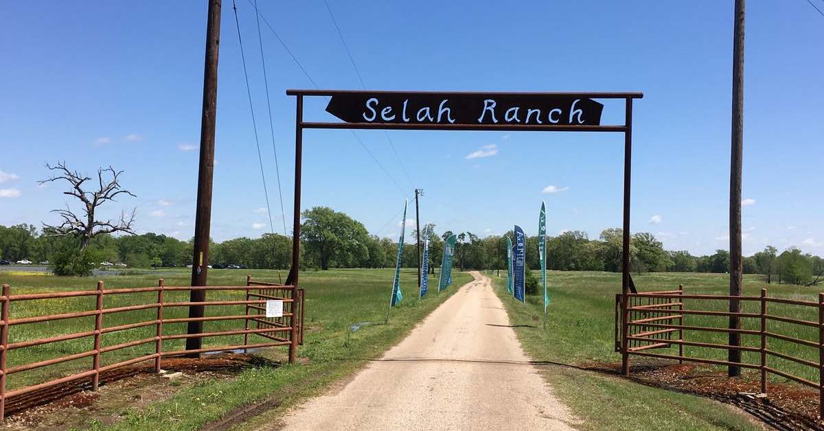 A dirt road through a flat, grassy Texas landscape with a metal sign over it saying "Selah Ranch"