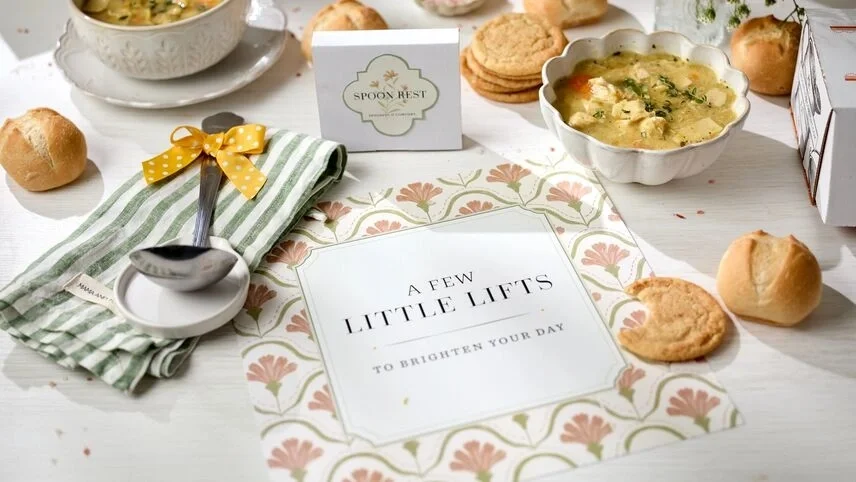 Spoonful of Comfort gift spread with tomato bisque, cookies, rolls, "A Few Little Lifts" card, and fresh flowers.