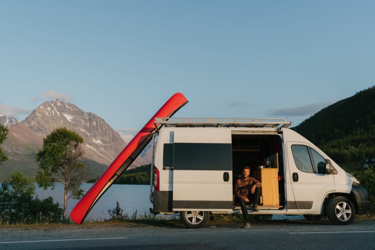 Man sitting in the doorframe of RV that's parked on the side of road with canoe leaning against van