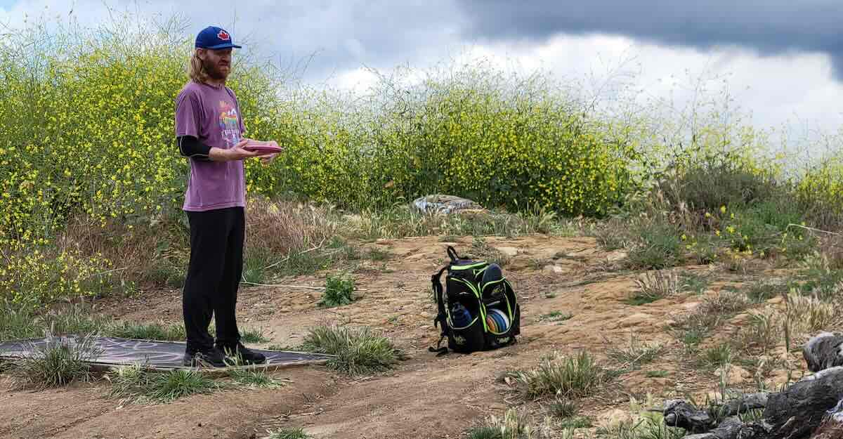 man in purple shirt and baseball cap on disc golf tee pad near yellow flowers