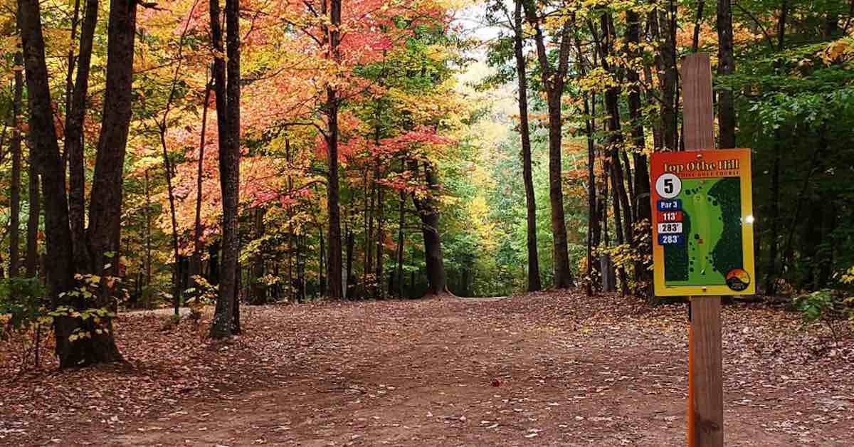 A tightly wooded disc golf fairway in fall colors. A tee sign is in the foreground in the right of the photo