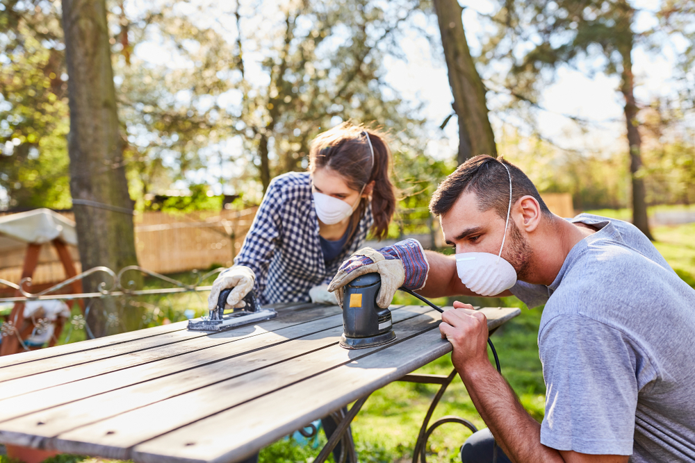 Couple wearing dust masks sand down a wooden table outdoors