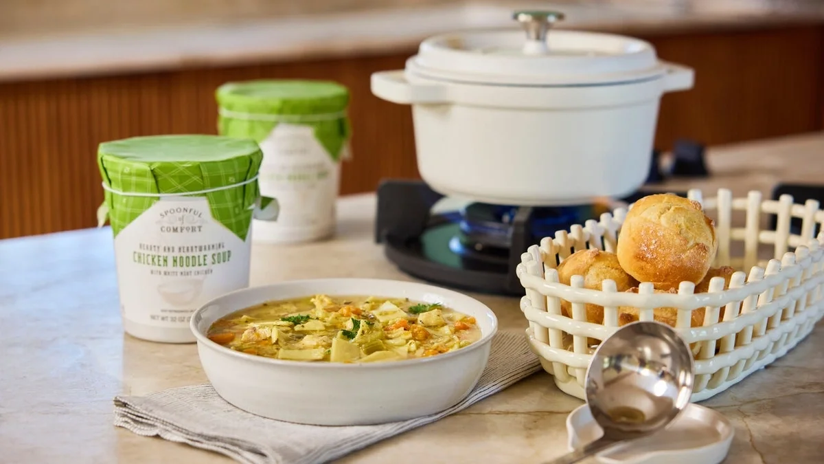 Bowl of chicken noodle soup served beside a basket of rolls, with two containers of chicken noodle soup, a metal ladle, and a Dutch oven on the stovetop in the background.
