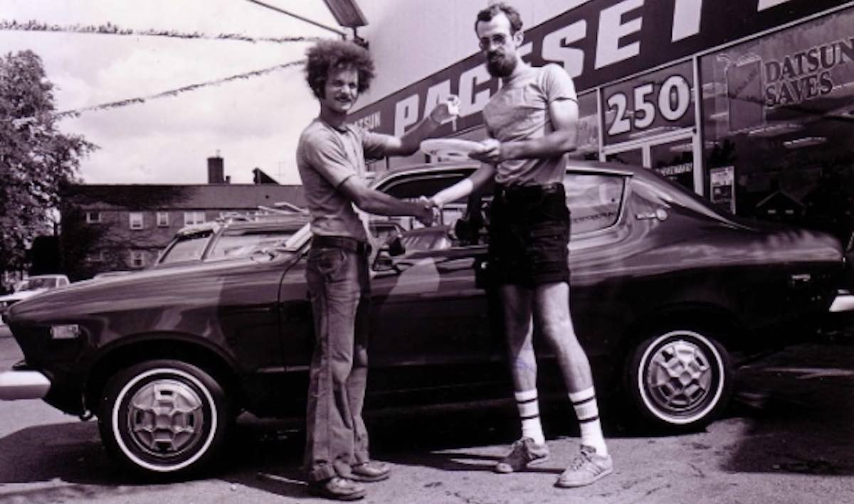 Two men stand in front of a new car in a black and white photo, one handing the keys to the other ceremonially