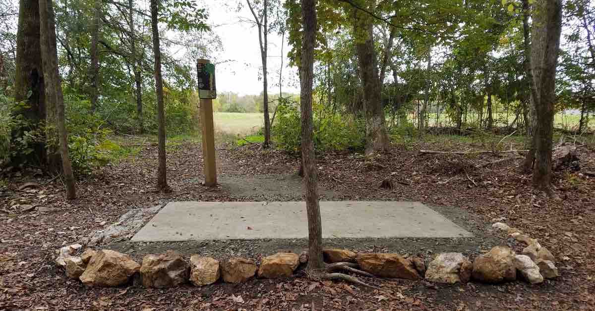 A concrete disc golf tee pad in a wooded area surrounded by rocks