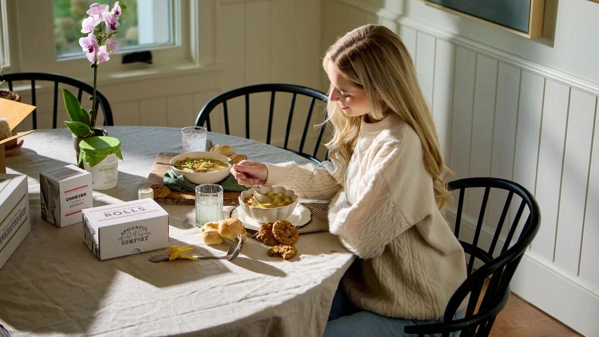 Woman sitting at a dining table enjoying a bowl of soup, with rolls, cookies, a glass of water, and Spoonful of Comfort gift boxes nearby.