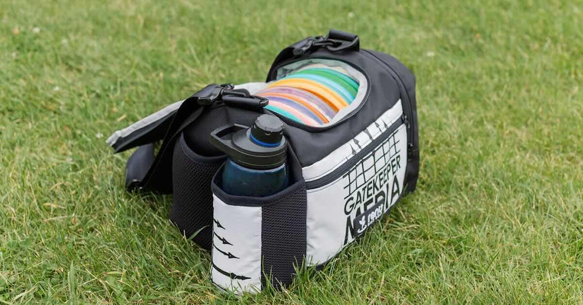 A gray and black single-strap disc golf bag with a water bottle and discs in it on the grass