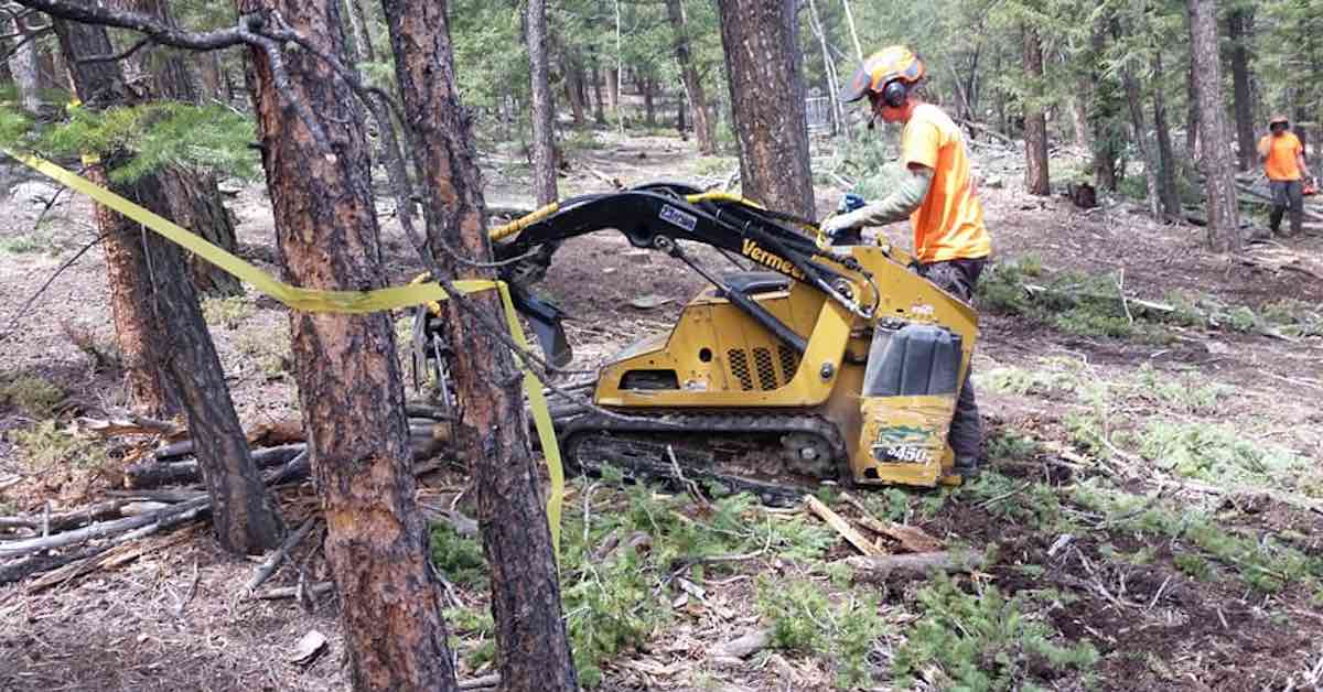 A man in an orange shirt and with ear protection on uses a large machine in the woods
