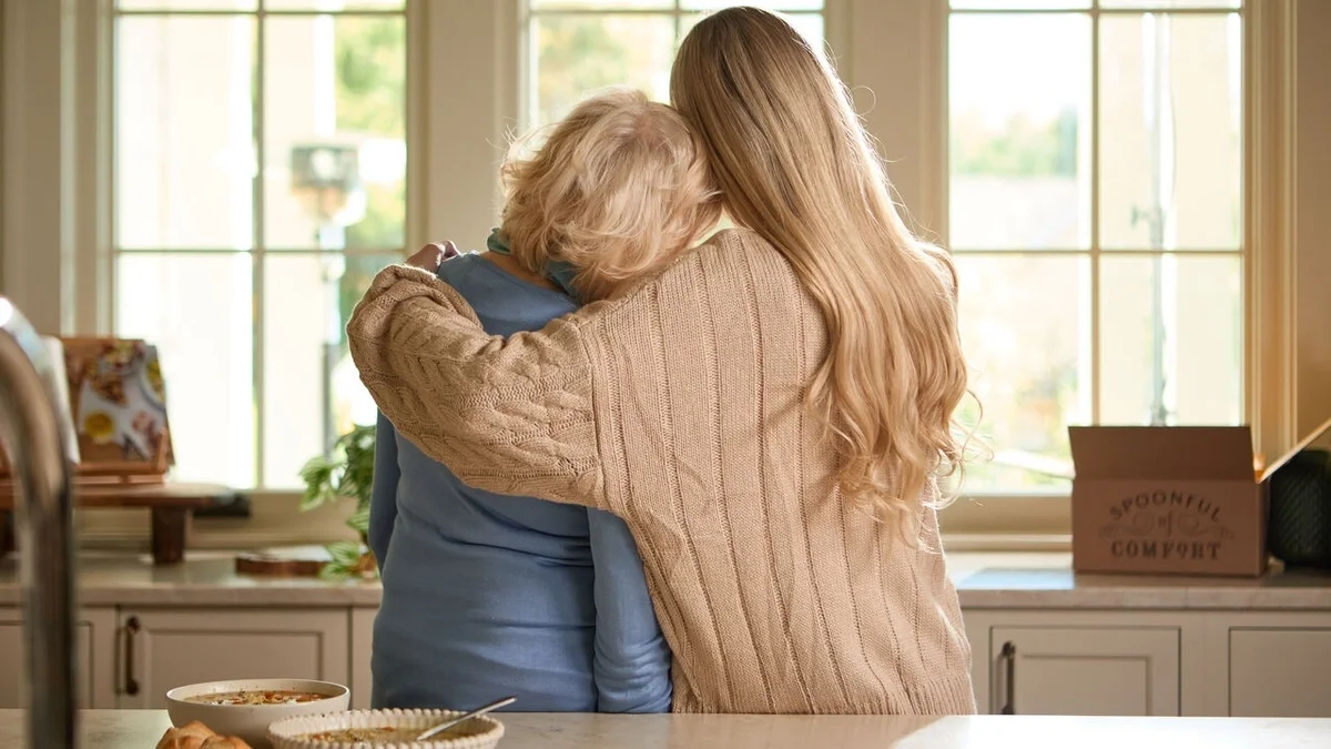Two people embracing in a bright kitchen, with bowls of soup on the counter and a Spoonful of Comfort gift box in the background.