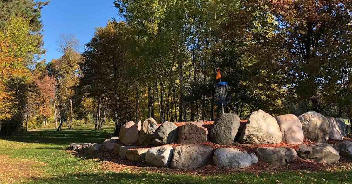 A blue disc golf basket on a large elevated green covered in red mulch and surrounded by boulders