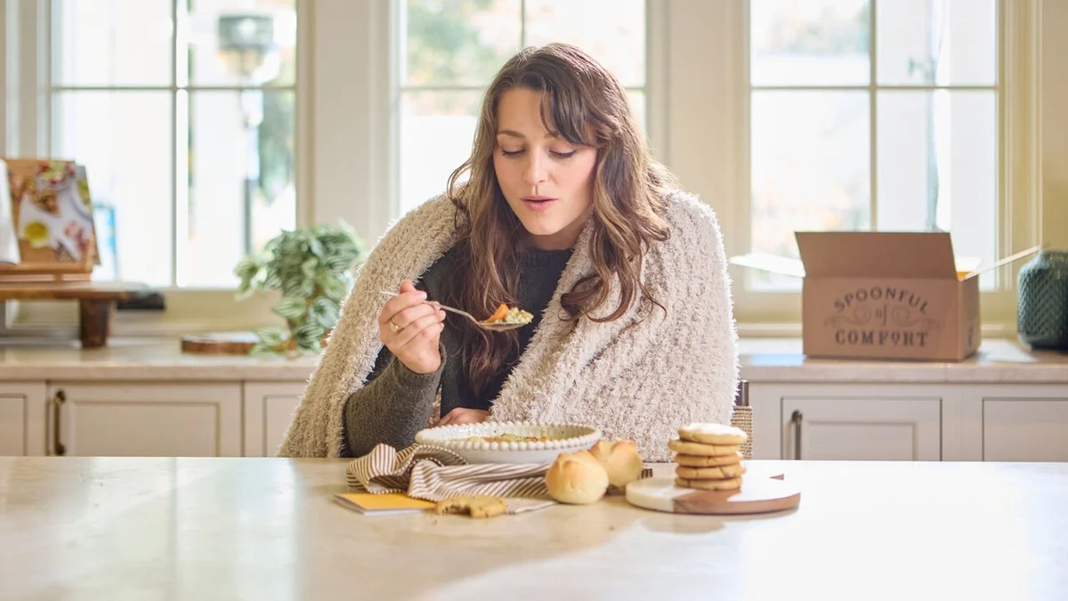 Woman wrapped in a cozy throw blanket, enjoying a bowl of soup at a kitchen counter with fresh rolls, cookies, and a Spoonful of Comfort gift box nearby.