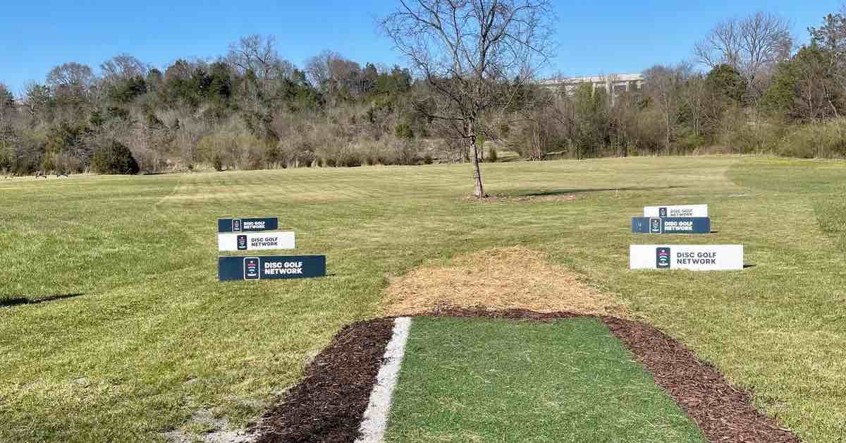 A turf disc golf tee in a mown area. DGPT signs in front of the tee in a line on the gorund