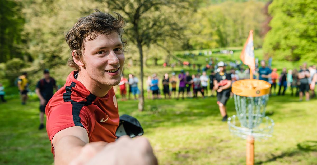 Young man fist bumping person behind the camera in front of a disc golf basket