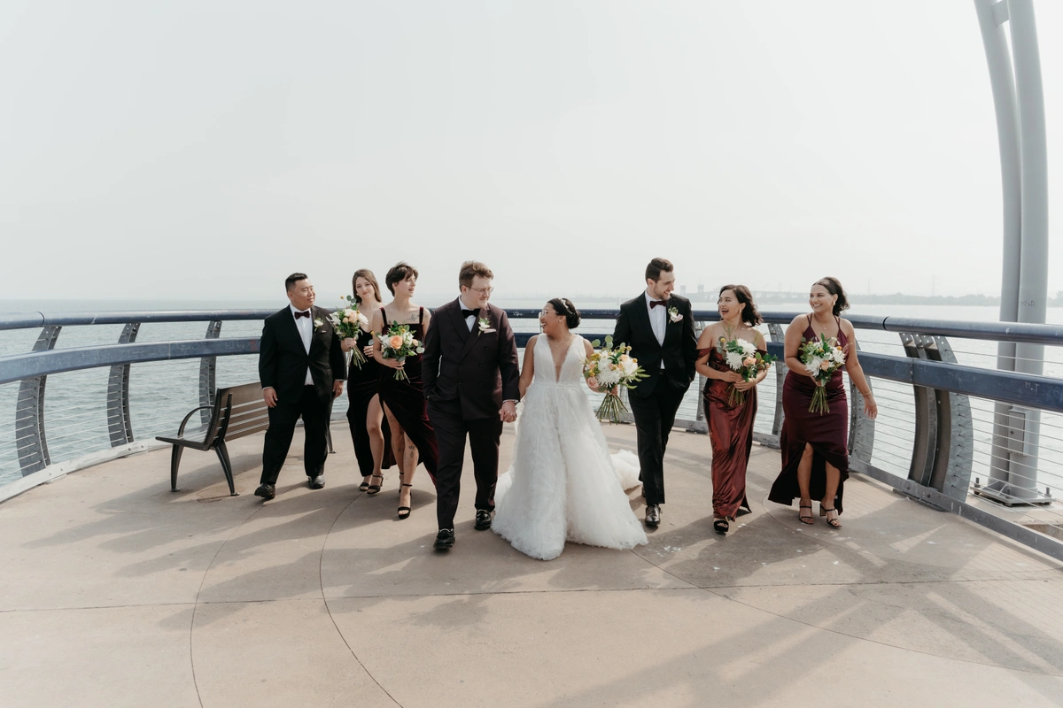 Bride and groom, with their wedding party, holding wedding flowers on a pier with water in the background.