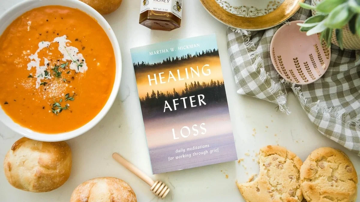 Overhead view of a sympathy gift arrangement featuring a bowl of creamy tomato soup, the book Healing After Loss, rolls, cookies, raw honey with a wooden dipper, and chamomile tea.