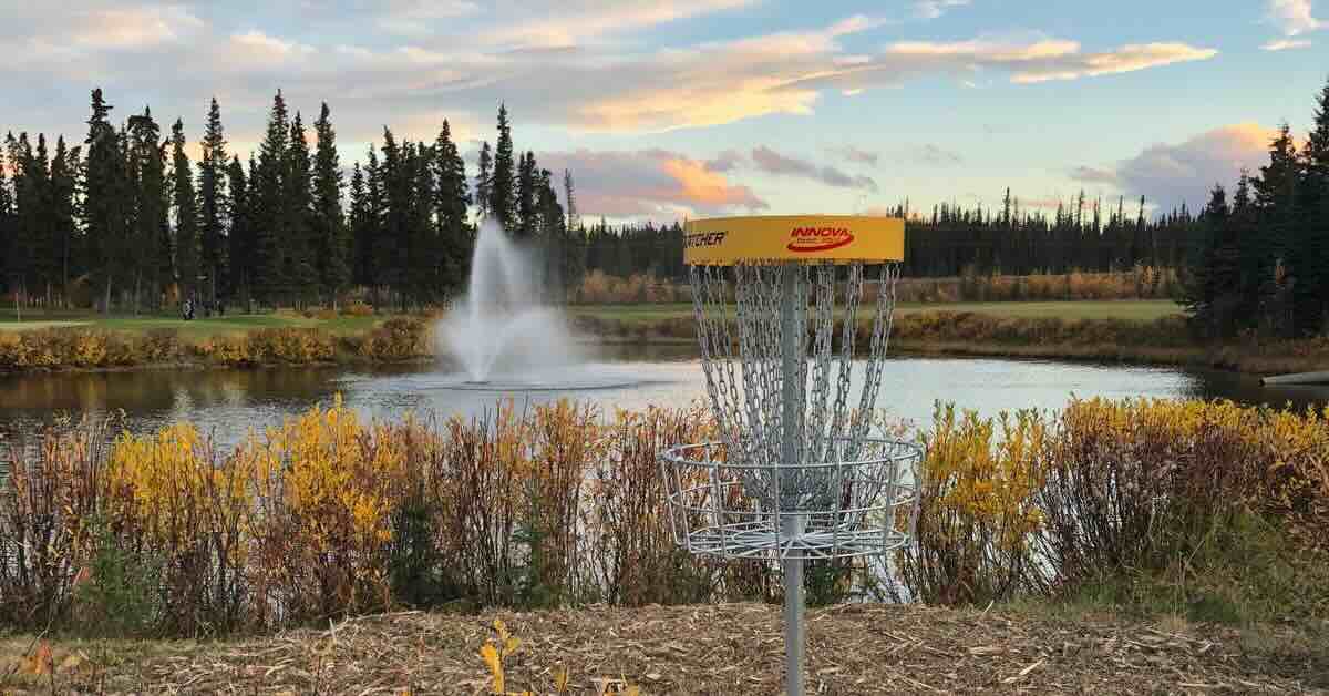 A disc golf basket in front of a pond with fountain and trees surrounding