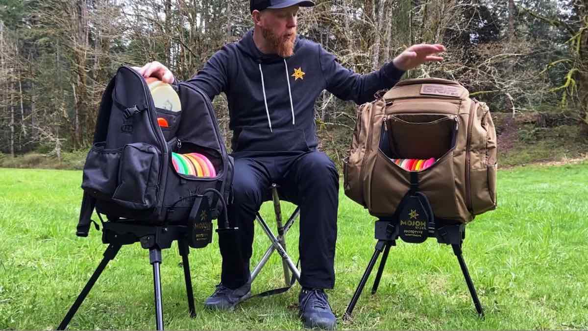 Man sitting on an outdoor stool between two disc golf bags on plastic, tripod stands