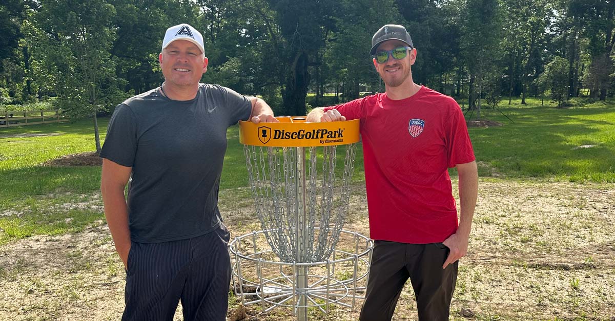 Two men in ball caps and tee shirts on either side of a disc golf basket posed for a photo