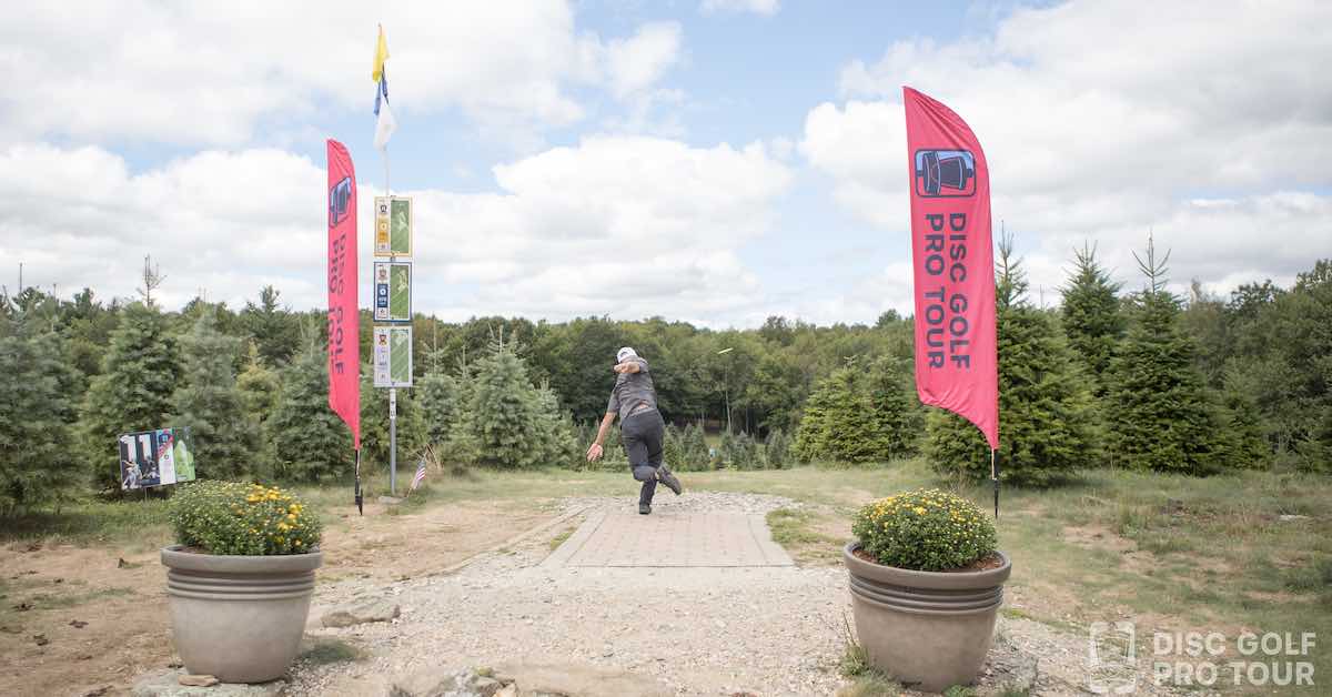 A view from far behind a tee pad with two large flower pots at its back edges. A person drives from the tee pad