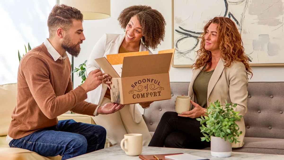 Three people sitting together in a room, smiling as they open a Spoonful of Comfort gift box, with mugs of coffee or tea on the table in front of them.