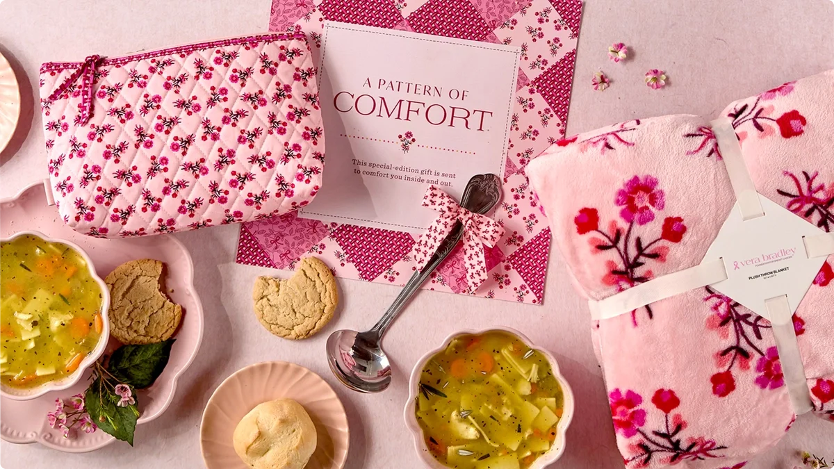 Close-up of a bowl of Tomato soup garnished with cream and herbs, surrounded by rolls, cookies, and a spoon on a pink serving board and Vera Bradley products, a blanket and cozy pouch.