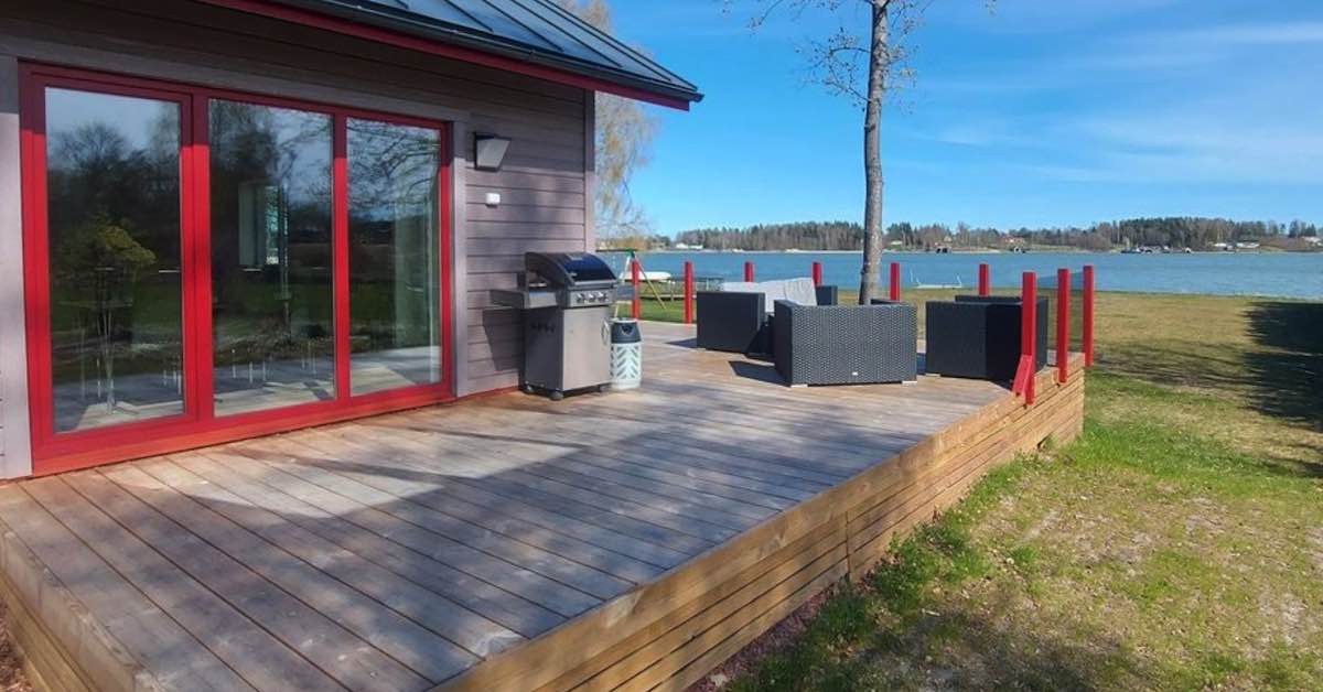 A deck with a view of the water and outside of a home painted gray with red window frames