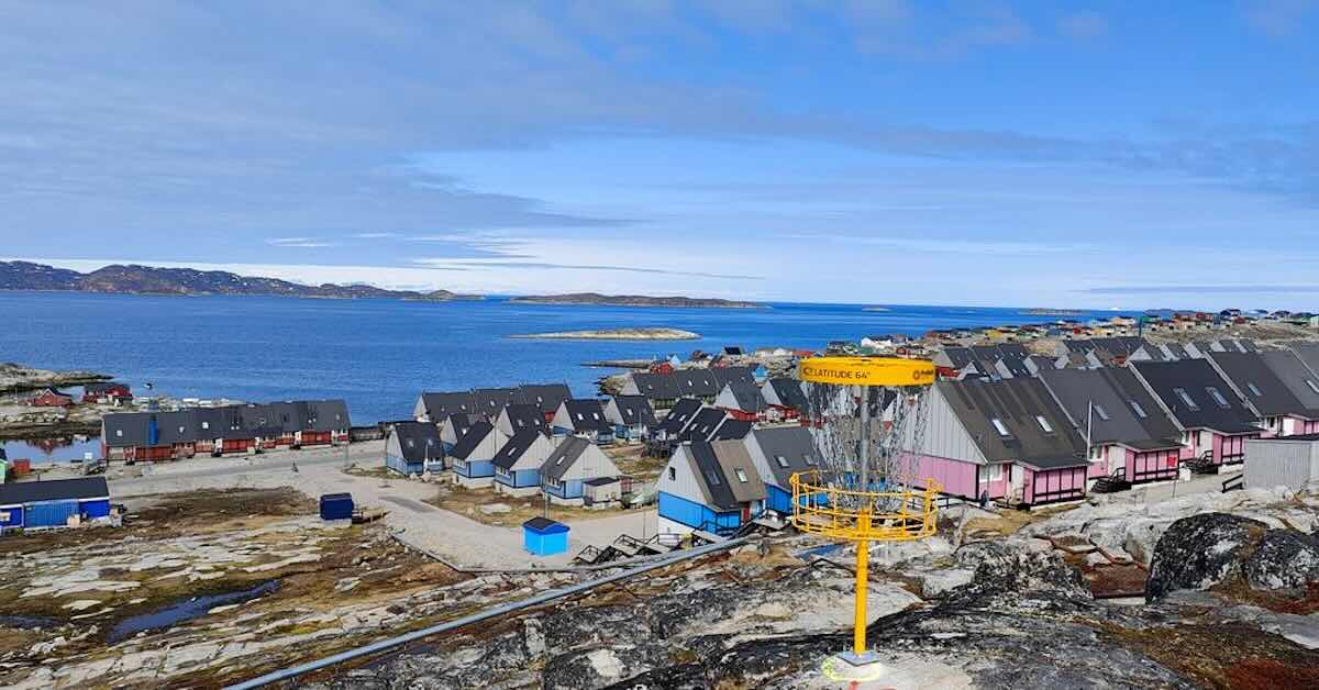 A yellow disc golf basket with a view of a village and sea in the background