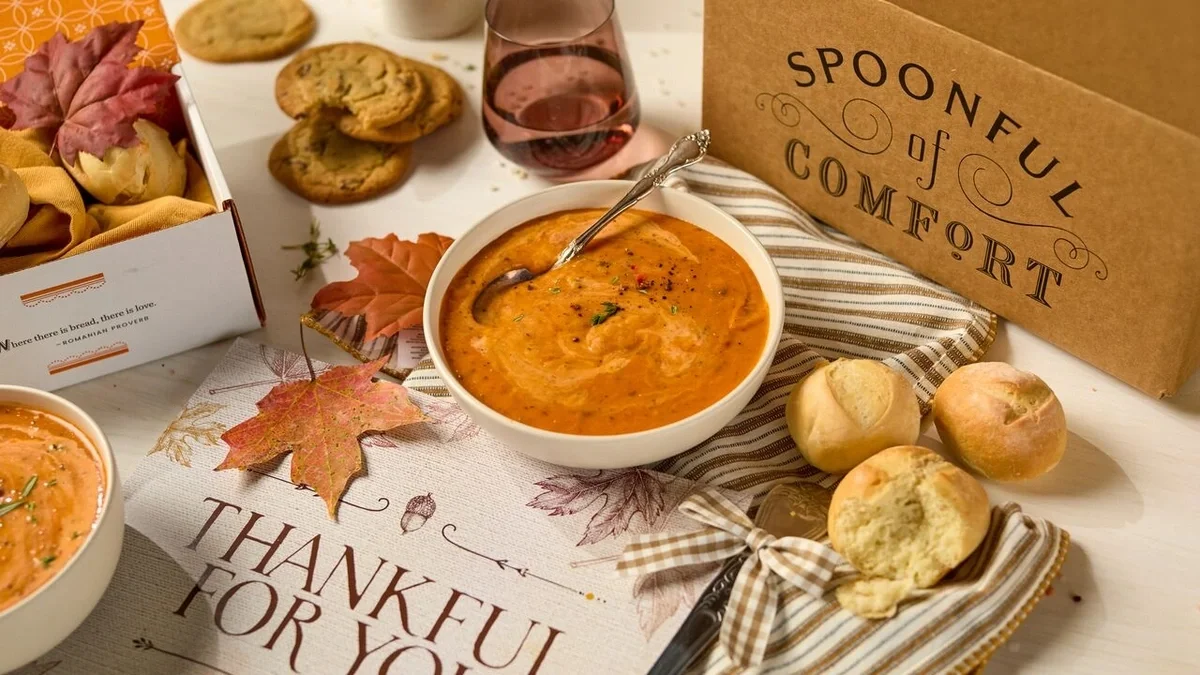 Fall-themed gift arrangement featuring a bowl of creamy soup, fresh rolls, cookies, autumn leaves, a glass of water, and a Spoonful of Comfort gift box displayed around a card that reads “Thankful for You.”