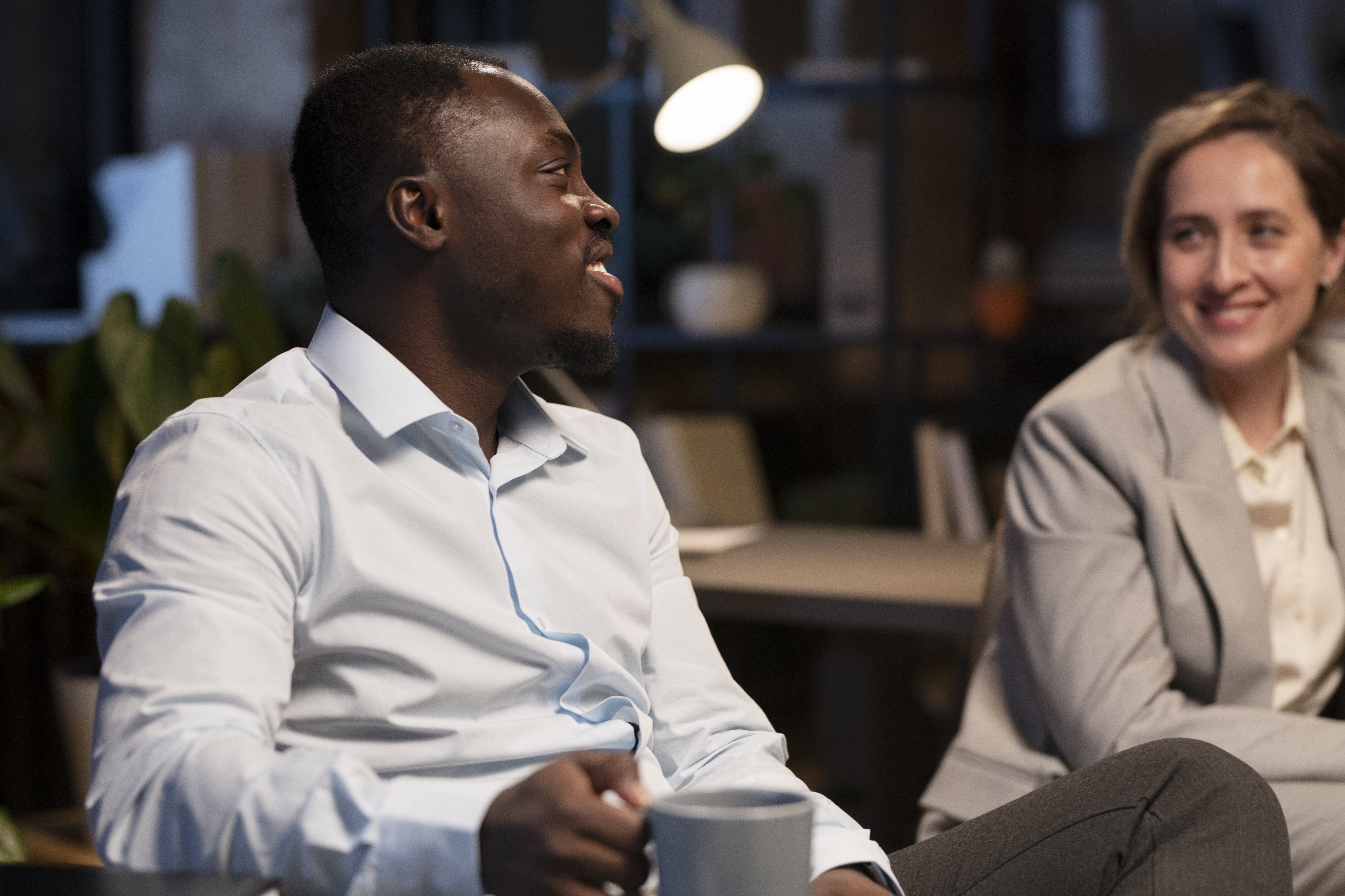 Two office workers having a friendly conversation. The man is smiling and holding a mug, while the woman, also smiling, listens attentively.