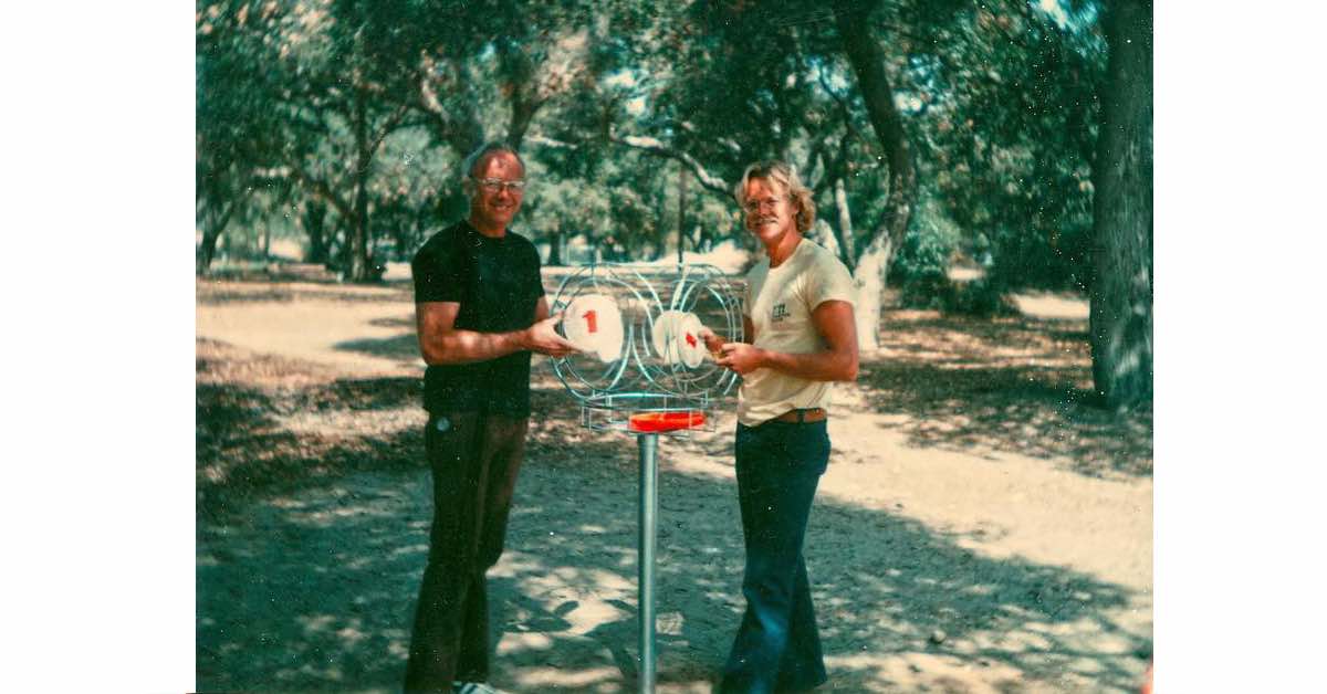 Two men in a park pose with an interesting disc catching device unlike modern disc golf baskets