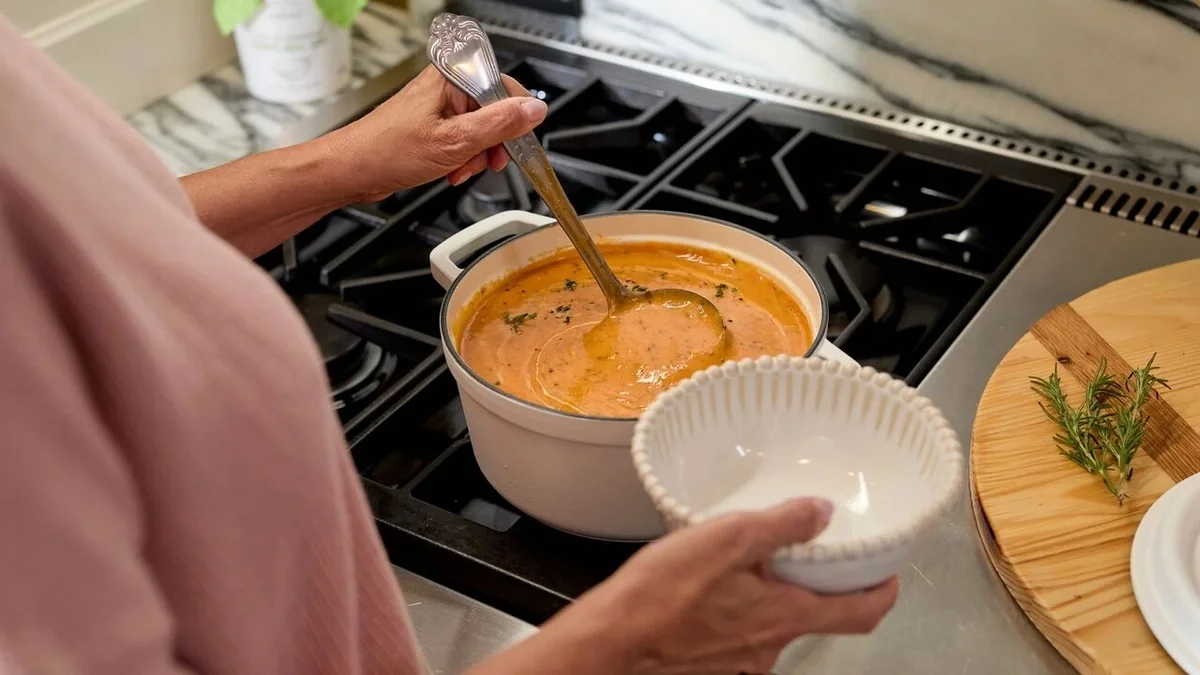 Person ladling creamy tomato soup from a white pot on the stovetop into a bowl, with fresh herbs and serving dishes nearby.