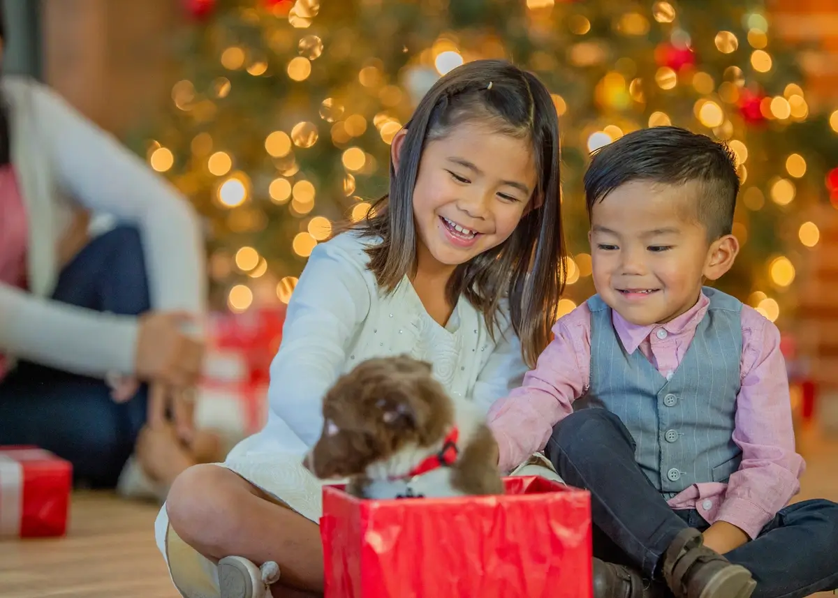 2 kids are happy to see a puppy jump out of a wrapped gift box at Christmas