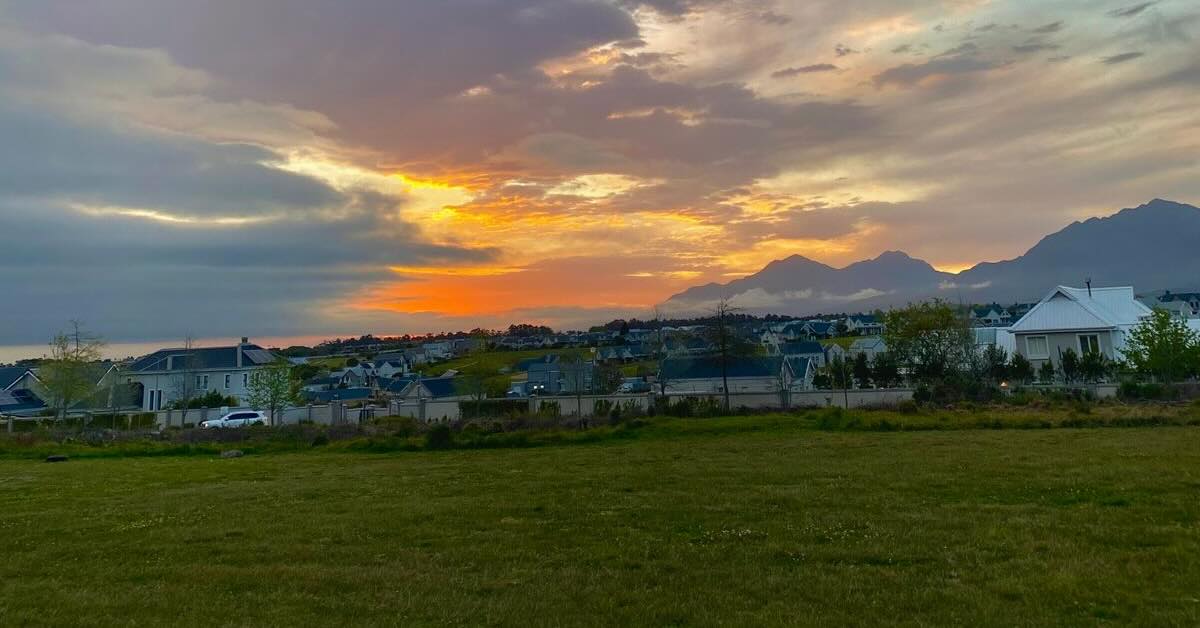 Open, green grass leads to a suburban neighborhood with a sunset and mountains in the distance