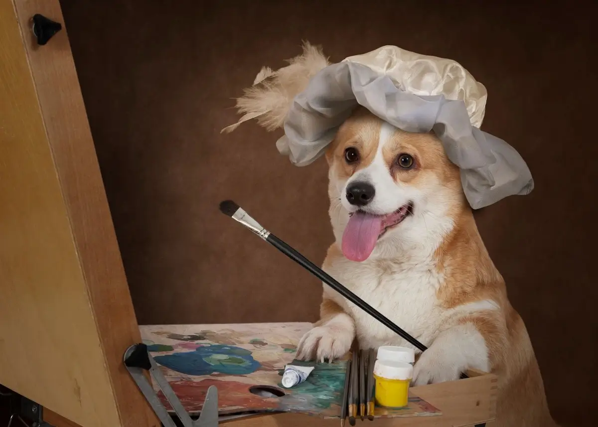 A dog wearing a frilly bonnet holds a paintbrush sitting across from an easel on a table
