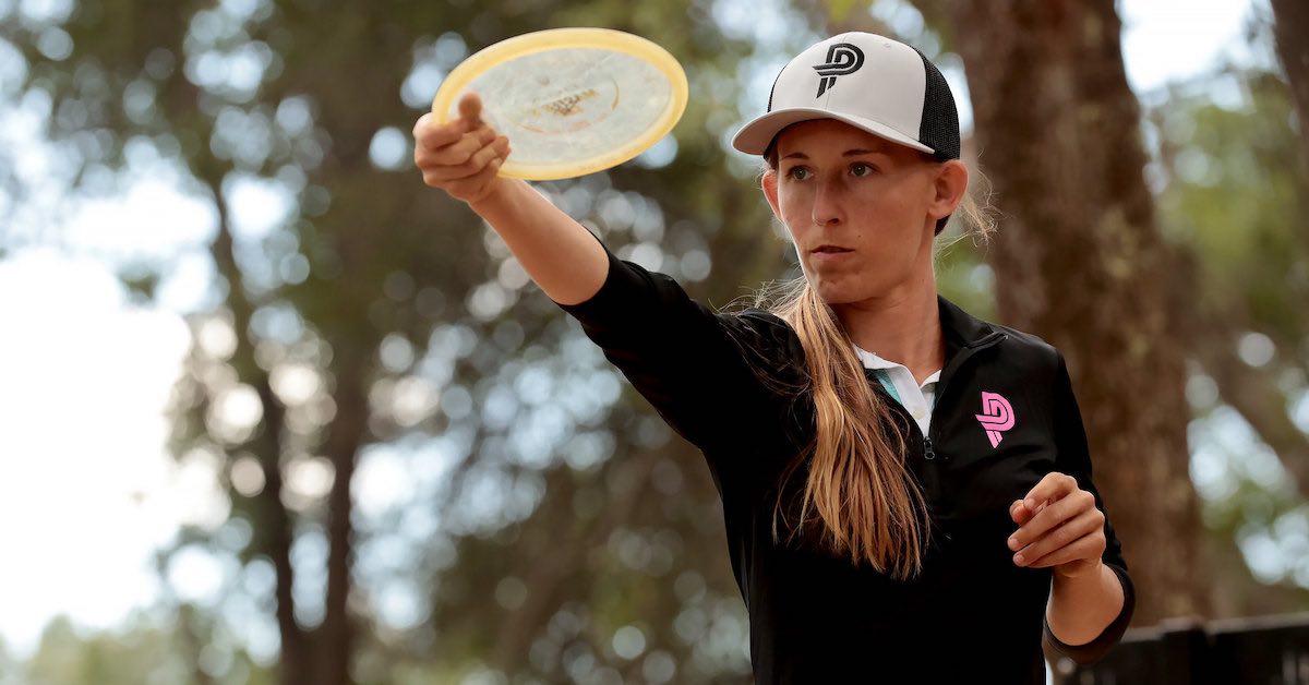 A young woman in a baseball cap holds a disc out, gauging a line to throw it on