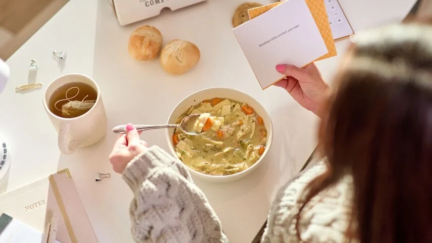 Overhead view of a person in a knit sweater eating a bowl of chicken soup while holding a handwritten note, with tea and rolls on the table nearby.