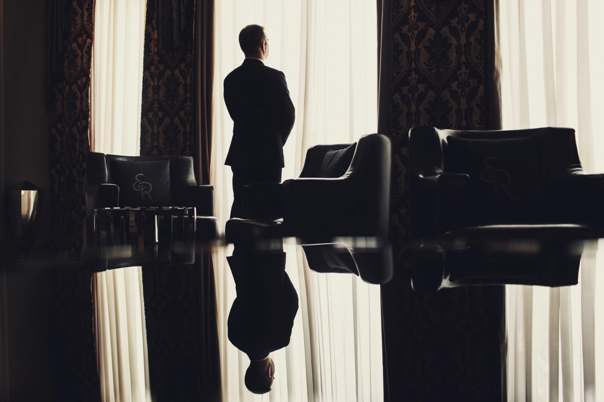 A man in formal attire stands silhouetted against a bright window, gazing outside. The room features luxurious furniture, including armchairs and a polished table reflecting his silhouette, creating an atmosphere of elegance and introspection.