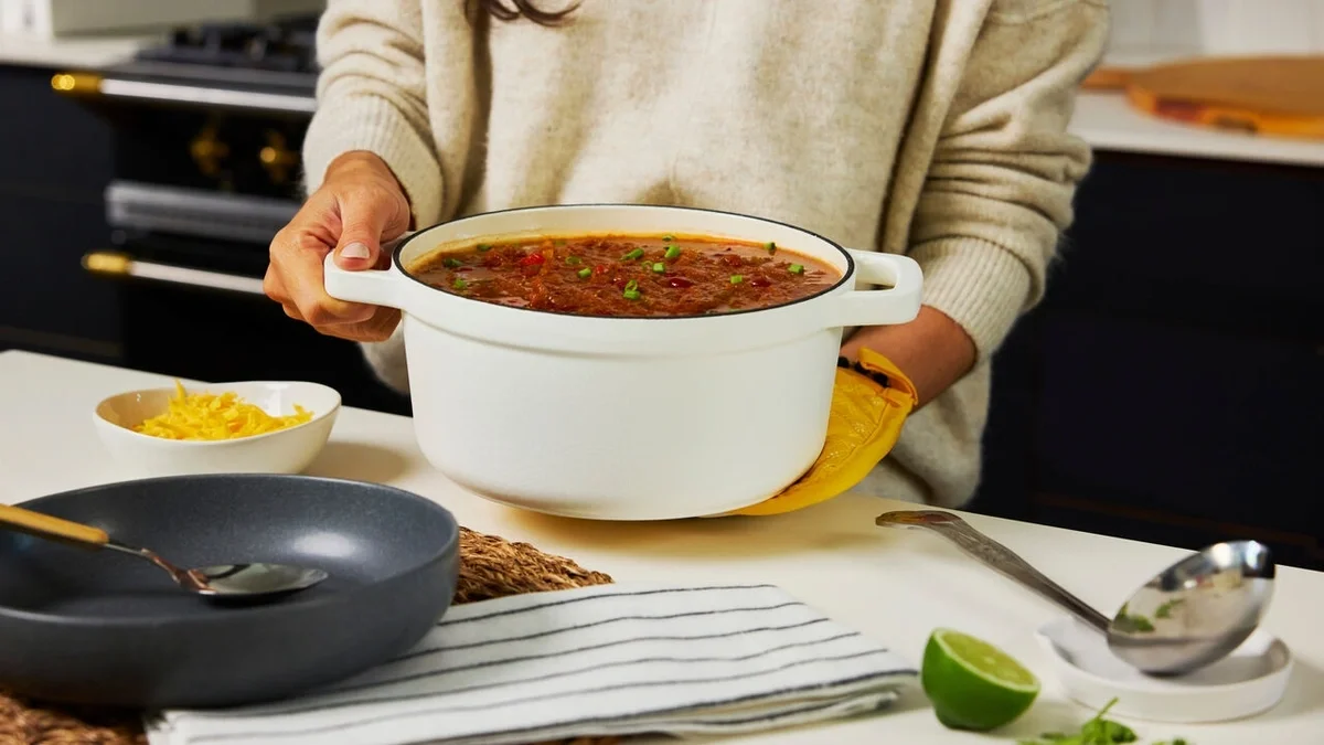 Person carrying a white Dutch oven filled with tomato soup, with shredded cheese, a skillet, a lime wedge, and serving utensils on the kitchen counter.