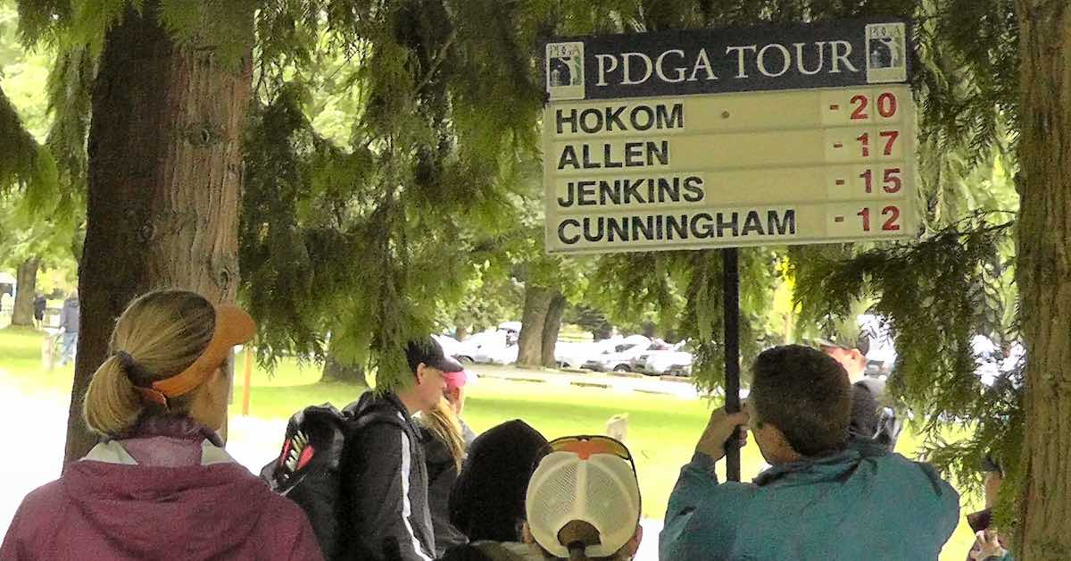 A crowd under evergreens seen from behind, one person holding a disc golf scoreboard