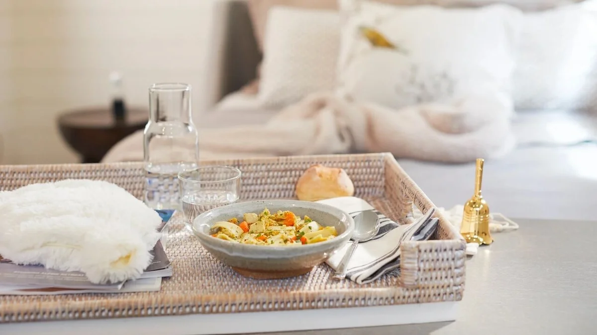 Tray arranged with a bowl of chicken soup, a roll, a glass carafe of water, silverware, a cozy throw blanket, and a gold bedside bell, with a neatly made bed in the background.