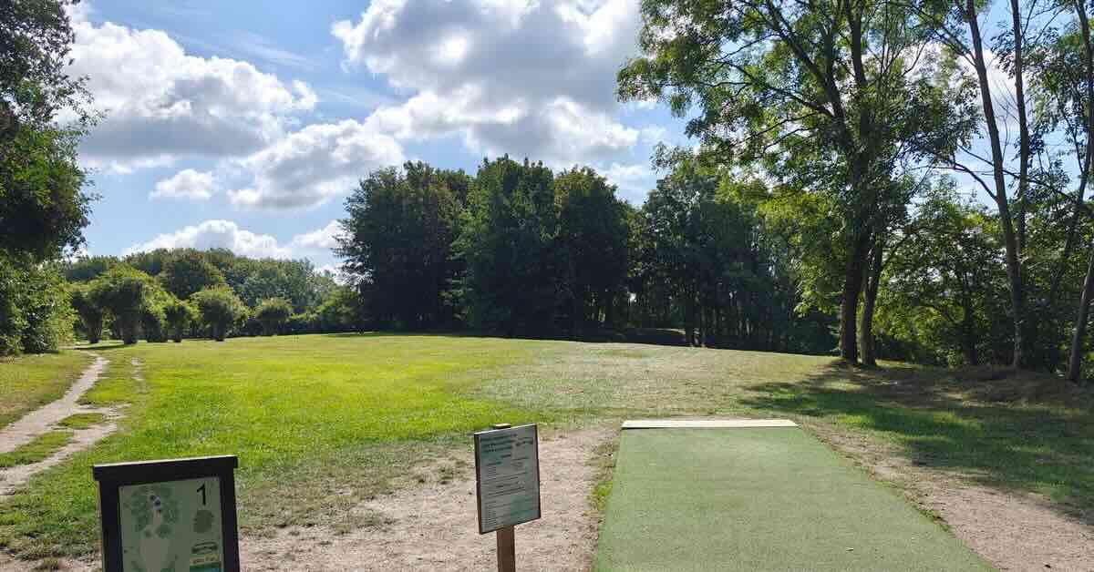 A turf disc golf tee pad in an open area leads to a green surround by trees