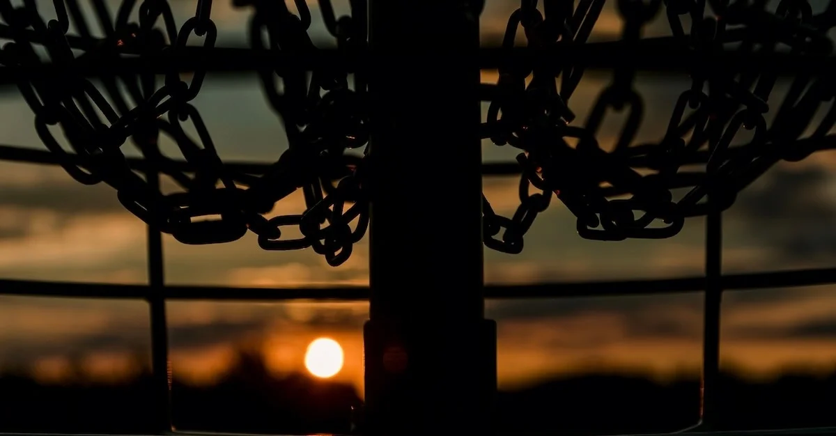 A setting sun seen through the shadowed basket and bottom of dangling chains in a disc golf basket