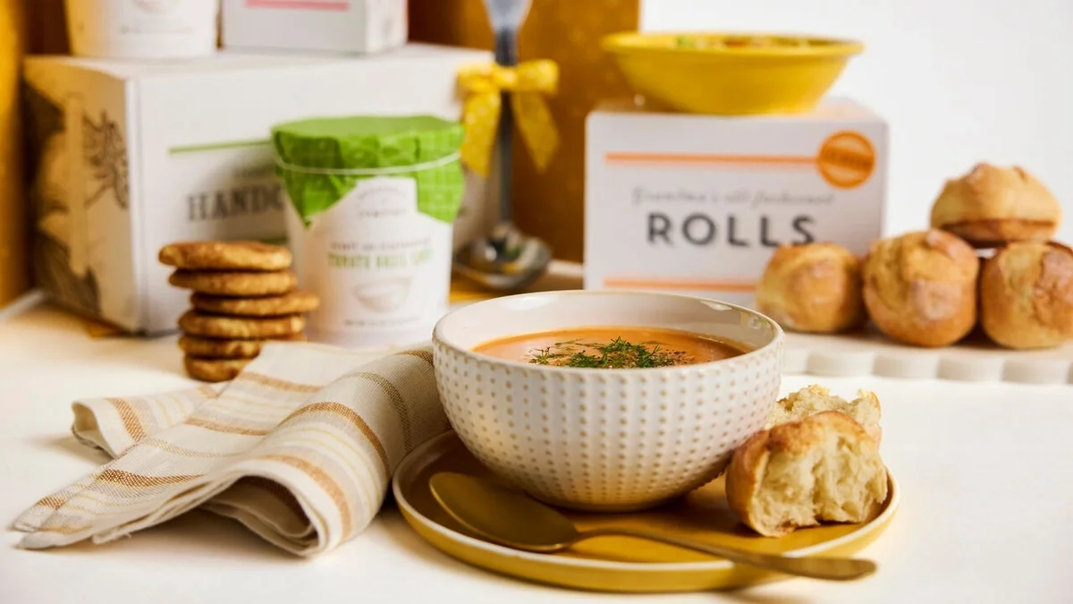 Bowl of creamy tomato soup served with a fresh roll, surrounded by rolls, cookies, packaged soup, and a metal ladle tied with a yellow ribbon.