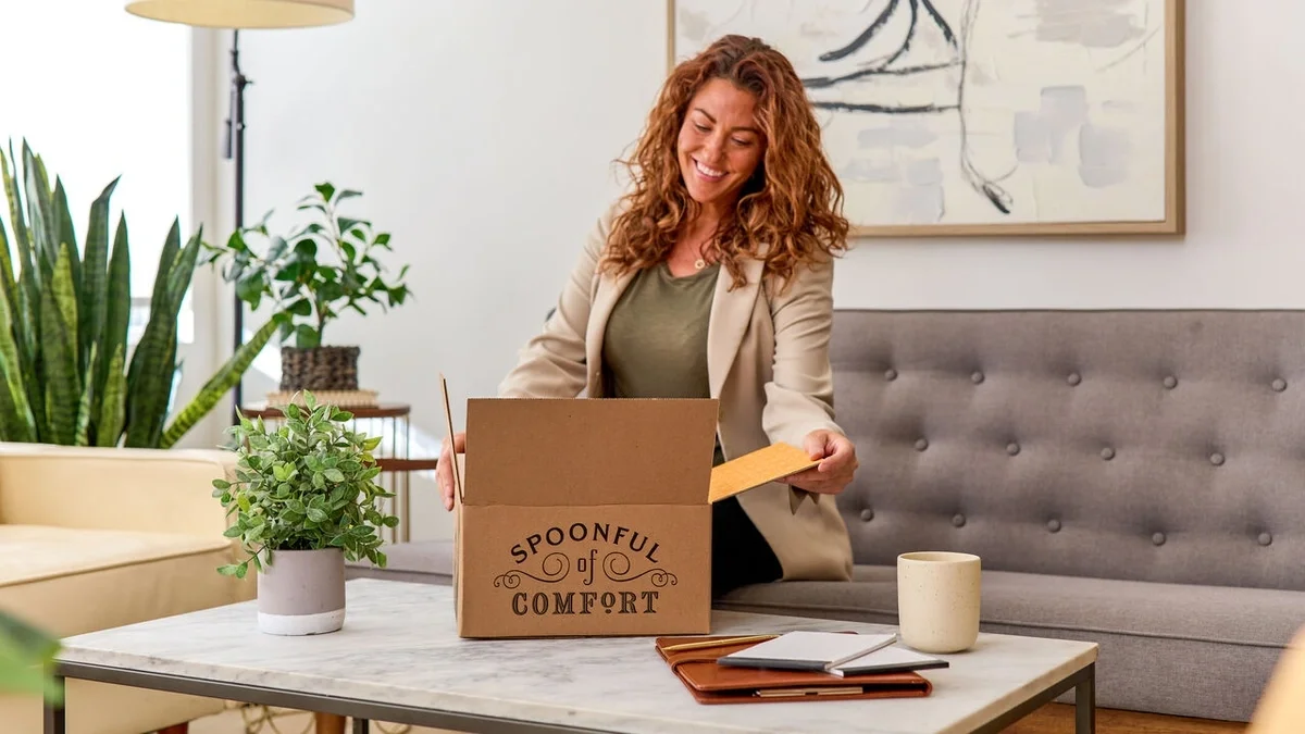 Woman smiling as she opens a Spoonful of Comfort gift box while sitting in a bright living room, with a coffee mug, notebook, and potted plants on the coffee table nearby.