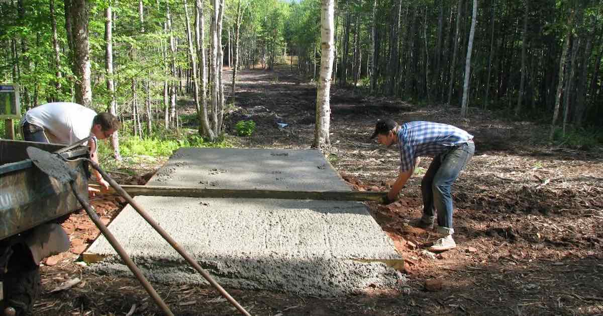 Two men use a board to level out wet concrete to make a disc golf tee pad