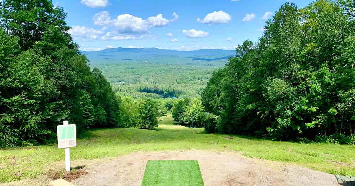 A disc golf tee pad looks down a grassy slope with very green trees on its sides and mountains in the distance