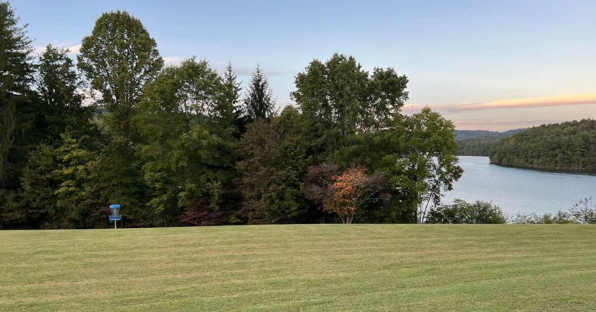 A wide swath of mown grass with a tree line, blue disc golf basket, and lake in the distance