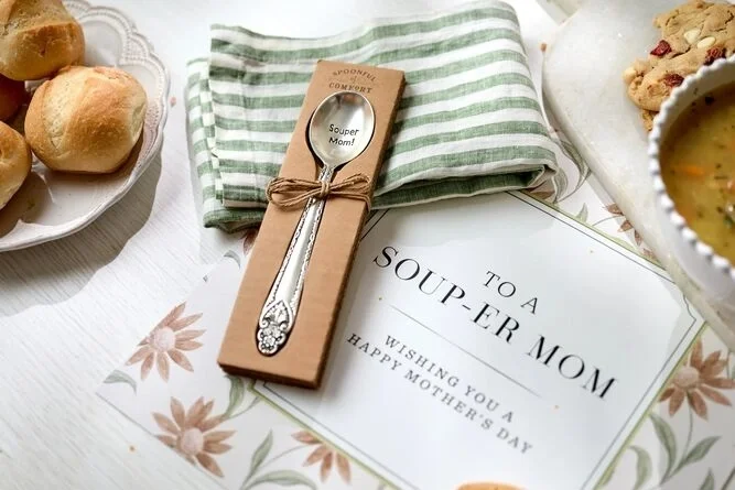 A close-up of a Mother’s Day place setting with a packaged spoon tied with twine, bread rolls on a plate, and a card that reads “To a Souper Mom — Wishing You a Happy Mother’s Day.”