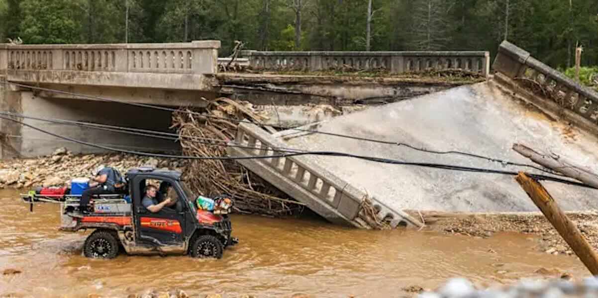 Men driving an ATV through a shallow river near a destroyed bridge