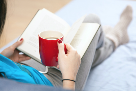 Image of woman from the neck down drinking a cup of tea and reading in bed 